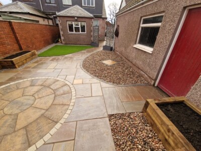 A stunning patio installation showcasing a unique combination of materials. A natural stone Indian sandstone patio serves as the foundation, exuding elegance and timeless charm. A Tobermore historic circle adds a touch of intricate design, creating a focal point within the space. Artificial grass is seamlessly integrated, providing a lush and low-maintenance area for relaxation and play. Timber sleepers border the patio, adding a rustic and organic element, while anthracite wall cladding brings a modern and sleek touch to the overall aesthetic. This patio installation harmoniously blends natural stone, artificial grass, timber sleepers, and anthracite wall cladding, resulting in a visually captivating and versatile outdoor space. Garden Landscaping Edinburgh, Garden Landscaping Alloa, Garden Rooms Edinburgh, Garden Rooms Alloa
