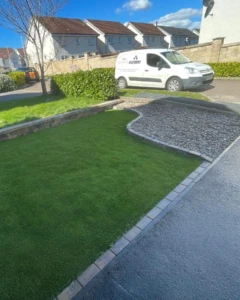 "Front garden design in Alloa featuring low-maintenance artificial grass and Scottish pebble stones. The boundary is defined by a Tobermore Bracken wall and Bracken kerbstone."