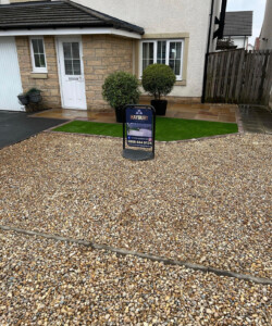 A stunning garden makeover in Alloa featuring an Indian Sandstone patio and decorative stones, creating a low-maintenance oasis. The lush green artificial grass replaces the traditional lawn, offering a hassle-free solution. The patio and decorative stones add an elegant touch to the overall design, providing a stylish and durable foundation. Transform your outdoor space with this beautiful and practical garden makeover." Driveway installation Edinburgh, Driveway Installation Alloa
